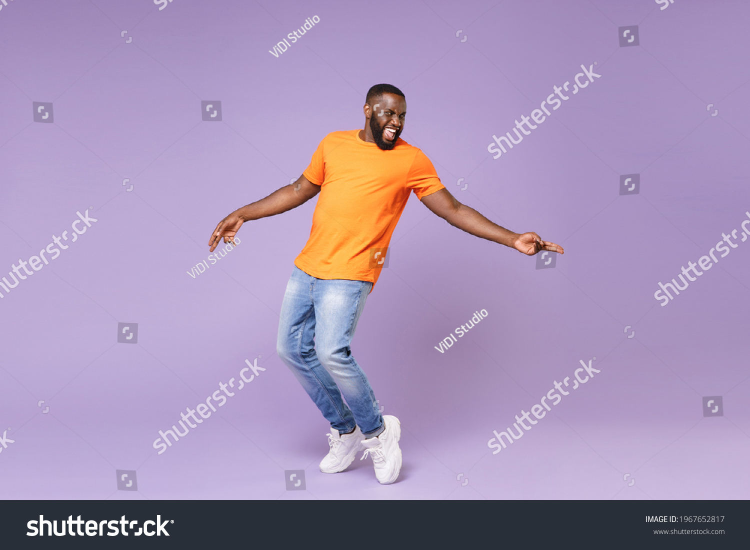 Volllange aufgeregter, lustiger junger afrikanischer Amerikaner der 20er Jahre in einfachem beigem orangefarbenem T-Shirt-Tanz, auf Zehen stehend, die Zeigefinger auf pastellviolettem Hintergrund, Studioportrait – Stockfoto